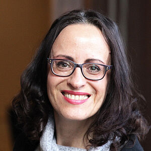 Portraitfoto von Iris Wemme-Baranowski. Sie blickt mit geöffnetem Mund lächelnd nach vorn. Ihr Haar ist an den Enden zu Locken geformt. Diese fallen ihr auf die Schulter. Sie trägt einen grau-melierten Pullover, dessen lockerer Rollkragen sich auf ihr Dekolleté legt. Darüber trägt sie einen schwarzen Cardigan.
