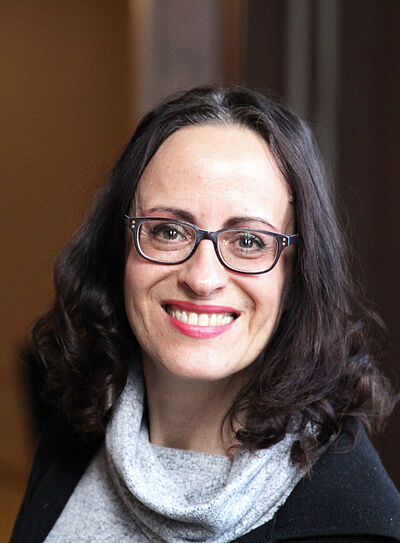 Portraitfoto von Iris Wemme-Baranowski. Sie blickt mit geöffnetem Mund lächelnd nach vorn. Ihr Haar ist an den Enden zu Locken geformt. Diese fallen ihr auf die Schulter. Sie trägt einen grau-melierten Pullover, dessen lockerer Rollkragen sich auf ihr Dekolleté legt. Darüber trägt sie einen schwarzen Cardigan.