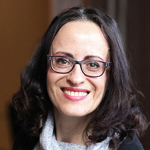 Portraitfoto von Iris Wemme-Baranowski. Sie blickt mit geöffnetem Mund lächelnd nach vorn. Ihr Haar ist an den Enden zu Locken geformt. Diese fallen ihr auf die Schulter. Sie trägt einen grau-melierten Pullover, dessen lockerer Rollkragen sich auf ihr Dekolleté legt. Darüber trägt sie einen schwarzen Cardigan.