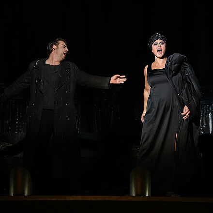 Calàf und Turandot stehen gemeinsam vorn auf der Bühne. Beide haben die Münder geöffnet und singen. Calàf strecht seine Hand nach Turandot aus. Sie schaut nach vorn, ihre Arme liegen an ihrem Körper.