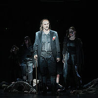 Macbeth steht in einen dunkelgrauen Anzug gekleidet mit einer Axt da. Seine Haare fallen ihm in Strähnen um das Gesicht. Am Boden liegen Menschen. Es laufen Hexen umher.