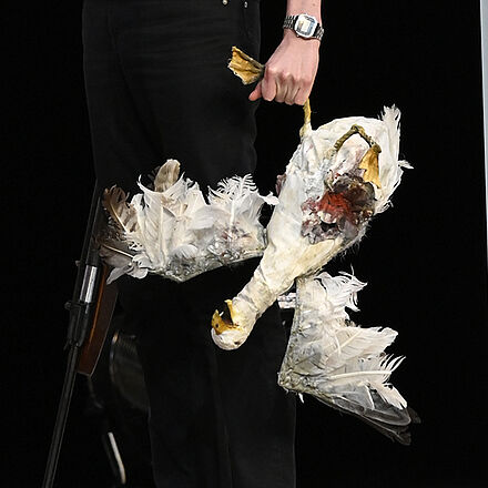 Ein Mann schaut zur Seite. Er steht an der Tür und hält eine tote Möwe in der rechten Hand. In der anderen Hand hält er eine Waffe.