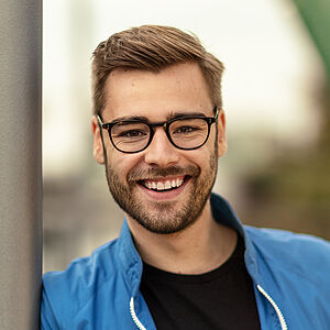 Portraitfoto von Luca Hämmerle. Er lehnt an einer grauen Stange und grinst mit geöffnetem Mund. Sein Haar ist braun, auf der rechten Seite gescheitelt und leicht nach oben gestylt. Er trägt eine blau-weiße Trainingsjacke, darunter ein schwarzes T-Shirt. Er trägt eine dunkle Brille und einen Dreitagebart.
