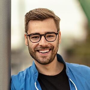 Portraitfoto von Luca Hämmerle. Er lehnt an einer grauen Stange und grinst mit geöffnetem Mund. Sein Haar ist braun, auf der rechten Seite gescheitelt und leicht nach oben gestylt. Er trägt eine blau-weiße Trainingsjacke, darunter ein schwarzes T-Shirt. Er trägt eine dunkle Brille und einen Dreitagebart.
