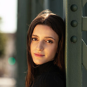 Portraitfoto von Giulia Girardi. Sie trägt ihr langes, braunes Haar offen und einen schwarzen Rollkragenpullover. Sie lehnt an einem grünen Metallstreben.