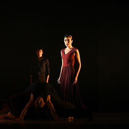 Eine Frau in Rot steht im Licht. Vor ihr liegen Tänzer:innen.