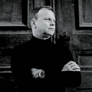 Schwarzweißes Portraitfoto von Stefan Vladar. Er steht mit verschränkten Armen vor einem großen Holzportal. Er trägt einen schwarzen Rollkragenpullover unter dem eine Armbanduhr hervorblitzt. Er blickt zur Seite.