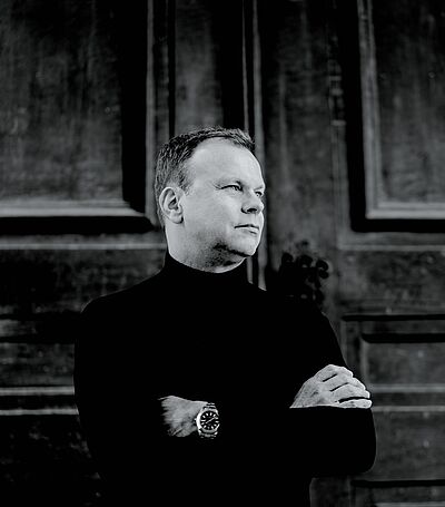 Schwarzweißes Portraitfoto von Stefan Vladar. Er steht mit verschränkten Armen vor einem großen Holzportal. Er trägt einen schwarzen Rollkragenpullover unter dem eine Armbanduhr hervorblitzt. Er blickt zur Seite.