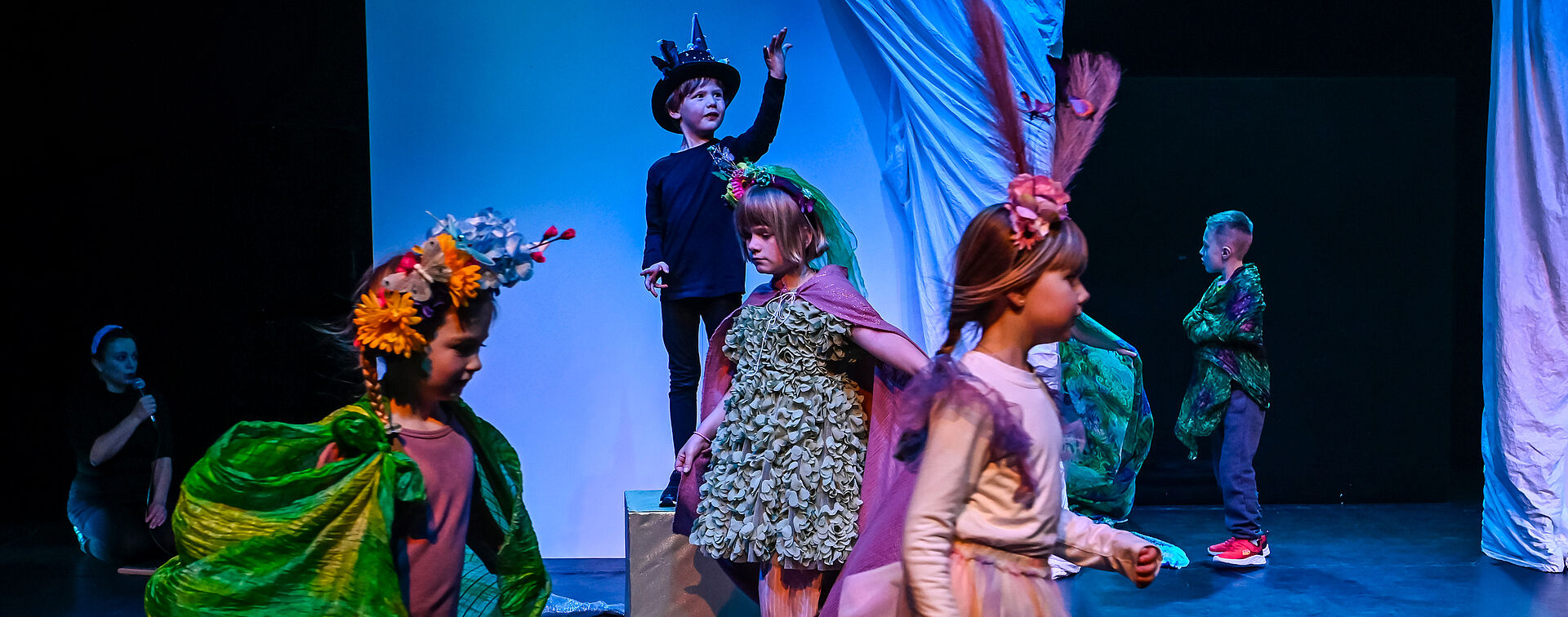 Kinder auf einer Bühne. Ein Mädchen steht mit einer Schneekugel im Vordergrund. Die Kinder dahinter hocken im Kreis oder auf goldenen Würfeln.