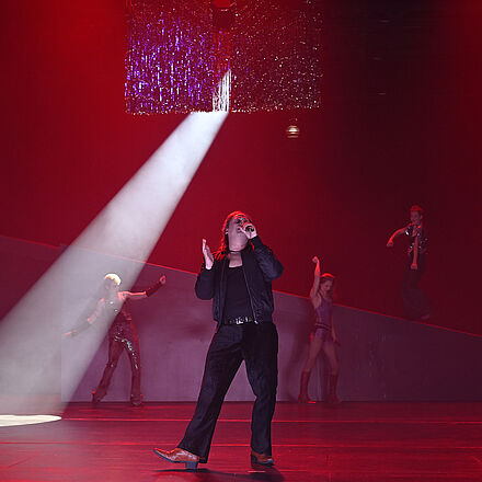 Im Vordergrund steht ein singender Mann, der ein Mikrofon in der Hand hält. Im Hintergrund tanzen drei Personen. Am Bildrand stehen zwei Grenzblockaden.