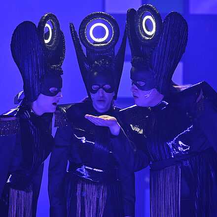 Im Mittelpunkt stehen drei gleich aussehende, schwarz gekleidete Personen. Die drei Personen schauen auf die Hand der rechten Person.