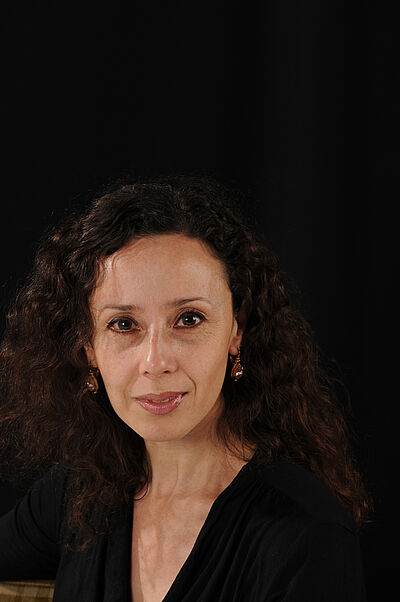 Portraitfoto von Adriana Mortelliti. Sie blickt leicht lächelnd in die Kamera. Ihr Körper ist seitlich gedreht. Sie trägt ihr lockiges dunkles Haar offen, dazu tränenförmige braune Ohrringe und ein schwarzes Oberteil mit V-Ausschnitt. Der Hintergrund ist schwar.