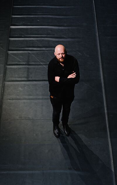 Bild von Florian Dragosits. Er steht mit verschränkten Armen aus schwarzem Bühnenboden. Er ist schwarz gekleidet und blickt nach oben in die Kamera. Sein Kopf ist rasiert, er trägt einen Vollbart.