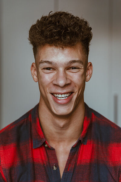 Portraitfoto von Malcolm Quinnten Henry. Er grinst mit geöffnetem Mund. Sein Haar trägt er kurz und oben lockig. Er trägt ein rot und schwarz kariertes Hemd.