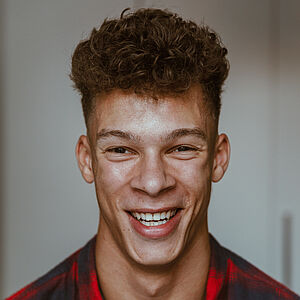 Portraitfoto von Malcolm Quinnten Henry. Er grinst mit geöffnetem Mund. Sein Haar trägt er kurz und oben lockig. Er trägt ein rot und schwarz kariertes Hemd.