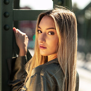 Portraitfoto von Mariagiovanna Fatima Bonavita. Sie ist vom Betrachter weggedreht und schaut über ihre linke Schulter. Ihr rechter Unterarm lehnt an einer grünen Stahlstrebe, mit der Hand hält sie sich daran fest. Ihr langes blondes Haar hängt wehend herunter. Sie trägt eine olivfarbene Samtbluse.