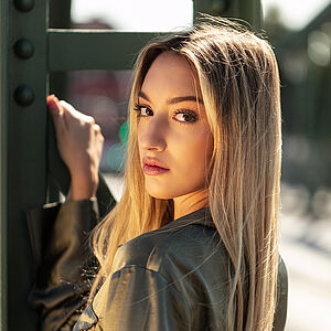 Portraitfoto von Mariagiovanna Fatima Bonavita. Sie ist vom Betrachter weggedreht und schaut über ihre linke Schulter. Ihr rechter Unterarm lehnt an einer grünen Stahlstrebe, mit der Hand hält sie sich daran fest. Ihr langes blondes Haar hängt wehend herunter. Sie trägt eine olivfarbene Samtbluse.