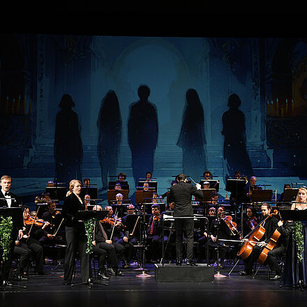 Im Vordergrund stehen vier Personen und singen, im Hintergrund ist das gesamte Orchester zu sehen. 