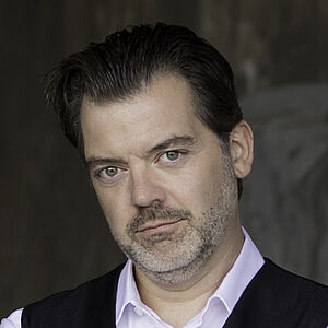 Portraitfoto von Patrick Ruyters. Sein Mund ist geschlossen, er lächelt dezent. Sein Kopf ist leicht schräg gelegt. Seine Haare sind zurückgekämmt. Er hat einen gräulichen Dreitagebart. Seine Arme sind vor der Brust verschränkt. Er trägt ein weißes Hemd und eine schwarze glänzende Bluse. Im Hintergrund ist verschwommen eine graue Wand zu erahnen.