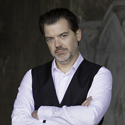 Portraitfoto von Patrick Ruyters. Sein Mund ist geschlossen, er lächelt dezent. Sein Kopf ist leicht schräg gelegt. Seine Haare sind zurückgekämmt. Er hat einen gräulichen Dreitagebart. Seine Arme sind vor der Brust verschränkt. Er trägt ein weißes Hemd und eine schwarze glänzende Bluse. Im Hintergrund ist verschwommen eine graue Wand zu erahnen.