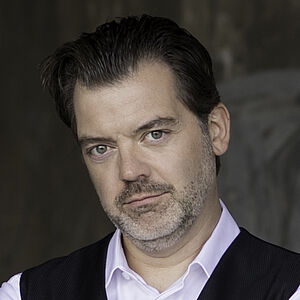 Portraitfoto von Patrick Ruyters. Sein Mund ist geschlossen, er lächelt dezent. Sein Kopf ist leicht schräg gelegt. Seine Haare sind zurückgekämmt. Er hat einen gräulichen Dreitagebart. Seine Arme sind vor der Brust verschränkt. Er trägt ein weißes Hemd und eine schwarze glänzende Bluse. Im Hintergrund ist verschwommen eine graue Wand zu erahnen.
