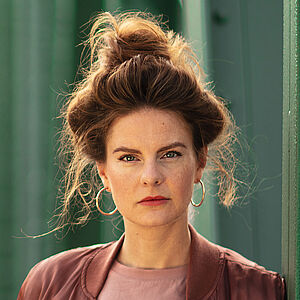 Portraitfoto von Severine Schabon. Sie lehnt seitlich an grünen Stahlstreben einer Brücke, ihre Hände sind in den Hosentaschen. Ihr Haar ist auf dem Kopf zu einem lockeren Dutt. Einzelne Strähnen hängen heraus. Sie trägt goldene Creolen, eine rotbraune Jacke, darunter ein Oberteil in altrosa, dazu eine hellgraue Jeans und einen schmalen beigefarbenen Gürtel mit silberner Schnalle.