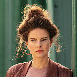 Portraitfoto von Severine Schabon. Sie lehnt seitlich an grünen Stahlstreben einer Brücke, ihre Hände sind in den Hosentaschen. Ihr Haar ist auf dem Kopf zu einem lockeren Dutt. Einzelne Strähnen hängen heraus. Sie trägt goldene Creolen, eine rotbraune Jacke, darunter ein Oberteil in altrosa, dazu eine hellgraue Jeans und einen schmalen beigefarbenen Gürtel mit silberner Schnalle.