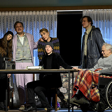 Eine Gruppe steht und sitzt am Tisch. Ein Mann schreibt an einer Schreibmaschine. Sie schauen in die Ferne.