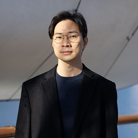 Portraitfoto von Jihaing Lee. Er ist in ein schwarzes Jackett gekleidet, darunter trägt er einen dunkelblauen Pullover. Im Gesicht trägt er eine dezente ovale Brille. Im Hintergrund ist ein weißes Segel.