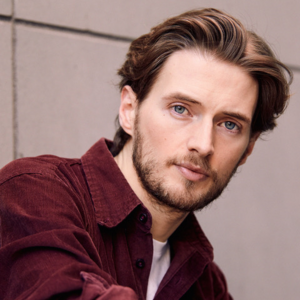 Portraitfoto von Tobias Joch. Er sitzt seitlich, der Kopf ist nach vorn gedreht. Er trägt ein rotes Hemd, darunter ein weißes T-Shirt.