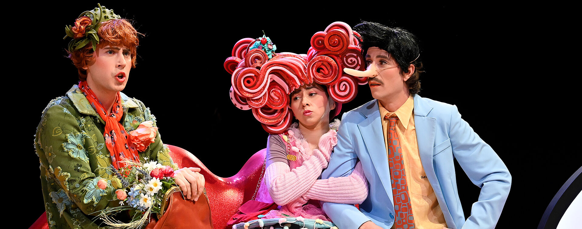 Drei Personen in unterschiedlichen Kostümen sitzen auf einer lippenförmigen Bank.