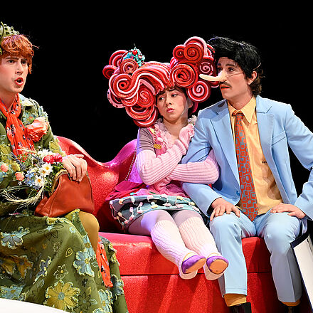 Drei Personen in unterschiedlichen Kostümen sitzen auf einer lippenförmigen Bank.