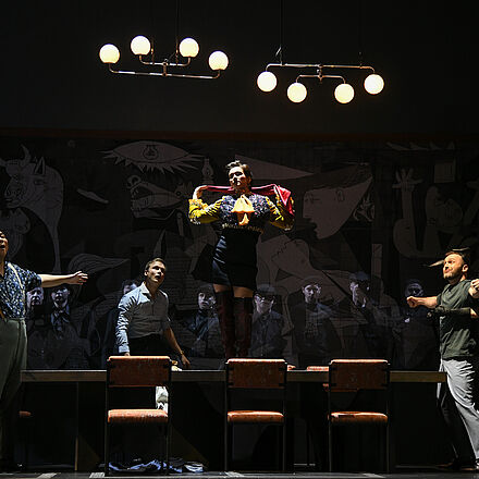 Im Mittelpunkt des Bildes steht ein Tisch, um den sich eine große Gruppe von Menschen versammelt hat. In der Mitte des Tisches steht eine Frau und hält einen Schal in den Händen. Alle Blicke sind auf sie gerichtet.