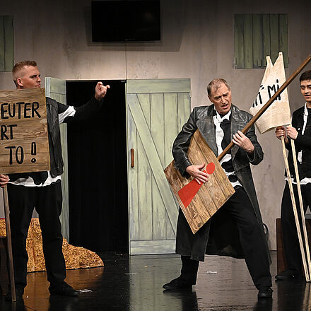 Vor einem Bauernhaus stehen drei Männer. Der ganz links hält ein Schild mit der Aufschrift: «MIEN EUTER HÖRT MI TO!» Rechts neben ihm spielt ein weiterer Mann mit einem Schild mit rotem Ausrufezeichen Luftgitarre. Rechts hinter ihm hält ein dritter, junger, Mann zwei weiße Fahnen, beide mit unlesbarer Aufschrift.