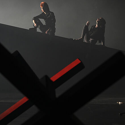 Im Hintergrund sitzen zwei Personen auf einer Erhöhung. Die rechte Person hält eine Zigarette in der Hand. Im Vordergrund ist eine unscharfe Grenzblockade zu sehen.
