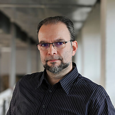 Portraitfoto von Róbert Tóth. Er steht leicht seitlich und blickt in die Kamera. Sein Haar ist zurückgekämmt. Er trägt eine eckige Brille und Bart. Er ist in ein schwarzes Hemd mit weißen Streifen gekleidet. Der Hintergrund ist verschwommen.