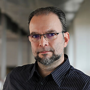 Portraitfoto von Róbert Tóth. Er steht leicht seitlich und blickt in die Kamera. Sein Haar ist zurückgekämmt. Er trägt eine eckige Brille und Bart. Er ist in ein schwarzes Hemd mit weißen Streifen gekleidet. Der Hintergrund ist verschwommen.