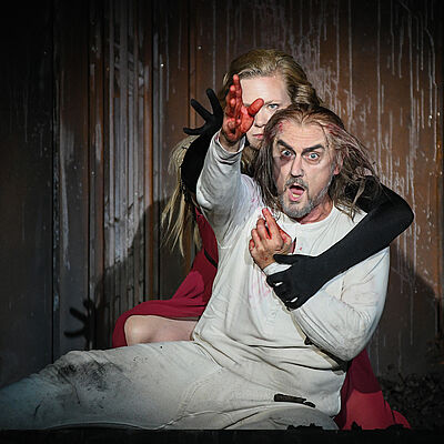 Lady Macbeth umgreift den am Boden sitzenden Macbeth von hinten. Ihre Arme sind in schwarze Handschuhe gehüllt. Er trägt weiße Unterwäsche und blickt mit weit aufgerissenen Augen nach vorn. Seine Hände sind blutverschmiert. Beide halten ihren rechten Arm empor. An der holzigen Rückwand laufen Spuren einer weißen Flüssigkeit herab.