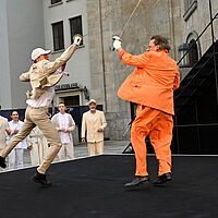 Fechtduell zwischen Cyrano und Valvert. Valvert, im beigen Anzug und bunter Krawatte, legt sich mit ganzer Kraft in den Angriff und springt auf seinen Kontrahenten Cyrano, im orangen Anzug, zu. Der Angegriffene pariert. Am Rand der Bühne stehen das Ensemble und feuert die Kämpfenden an.