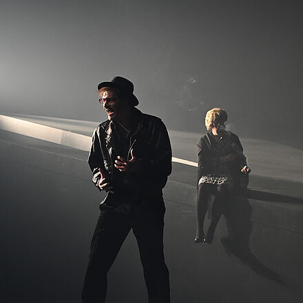 Im Vordergrund steht ein dunkel gekleideter Mann mit Hut. Im Hintergrund sitzt eine Person im Kleid auf einer Erhöhung.