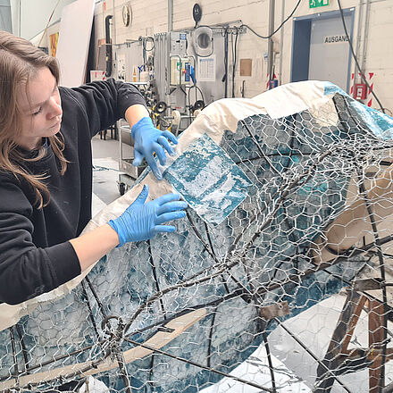 Der Drachenkopf wird mit in Holzleim getränkten Stofflappen beklebt. Die Arbeit findet im Malsaal statt.