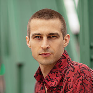 Portraitfoto von Richard Feist. Er schaut seitlich über die linke Schulter. Er trägt das Haar kurz geschoren und ein rot-schwarz gemustertes Hemd.