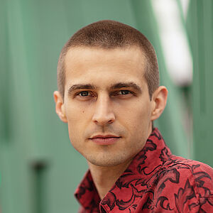 Portraitfoto von Richard Feist. Er schaut seitlich über die linke Schulter. Er trägt das Haar kurz geschoren und ein rot-schwarz gemustertes Hemd.