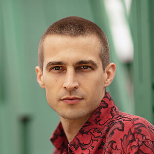Portraitfoto von Richard Feist. Er schaut seitlich über die linke Schulter. Er trägt das Haar kurz geschoren und ein rot-schwarz gemustertes Hemd.