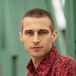 Portraitfoto von Richard Feist. Er schaut seitlich über die linke Schulter. Er trägt das Haar kurz geschoren und ein rot-schwarz gemustertes Hemd.
