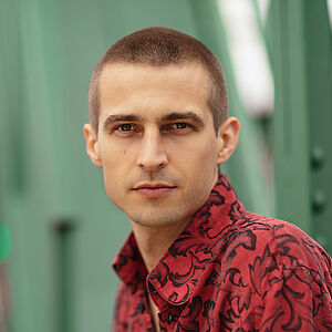 Portraitfoto von Richard Feist. Er schaut seitlich über die linke Schulter. Er trägt das Haar kurz geschoren und ein rot-schwarz gemustertes Hemd.