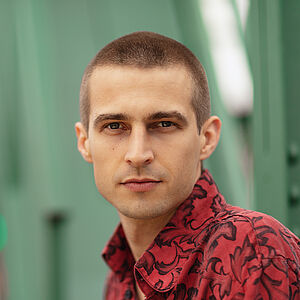 Portraitfoto von Richard Feist. Er schaut seitlich über die linke Schulter. Er trägt das Haar kurz geschoren und ein rot-schwarz gemustertes Hemd.