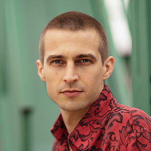 Portraitfoto von Richard Feist. Er schaut seitlich über die linke Schulter. Er trägt das Haar kurz geschoren und ein rot-schwarz gemustertes Hemd.