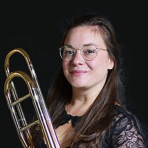 Portraitfoto von Camille Renaud. Sie steht in schwarzer Kulisse und hält eine Posaune in den Händen. Sie trägt ein schwarzes Kleid mit Spitze. Sie hat braunes langes Haar und trägt eine Brille. Sie lächelt in die Kamera.