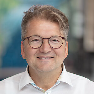 Portraitfoto von Lars Tietje. Er trägt eine runde Brille und ein weißes Hemd. Seine Hände stecken in den Hosentaschen. Er grinst leicht. Der Hintergrund ist verschwommen.
